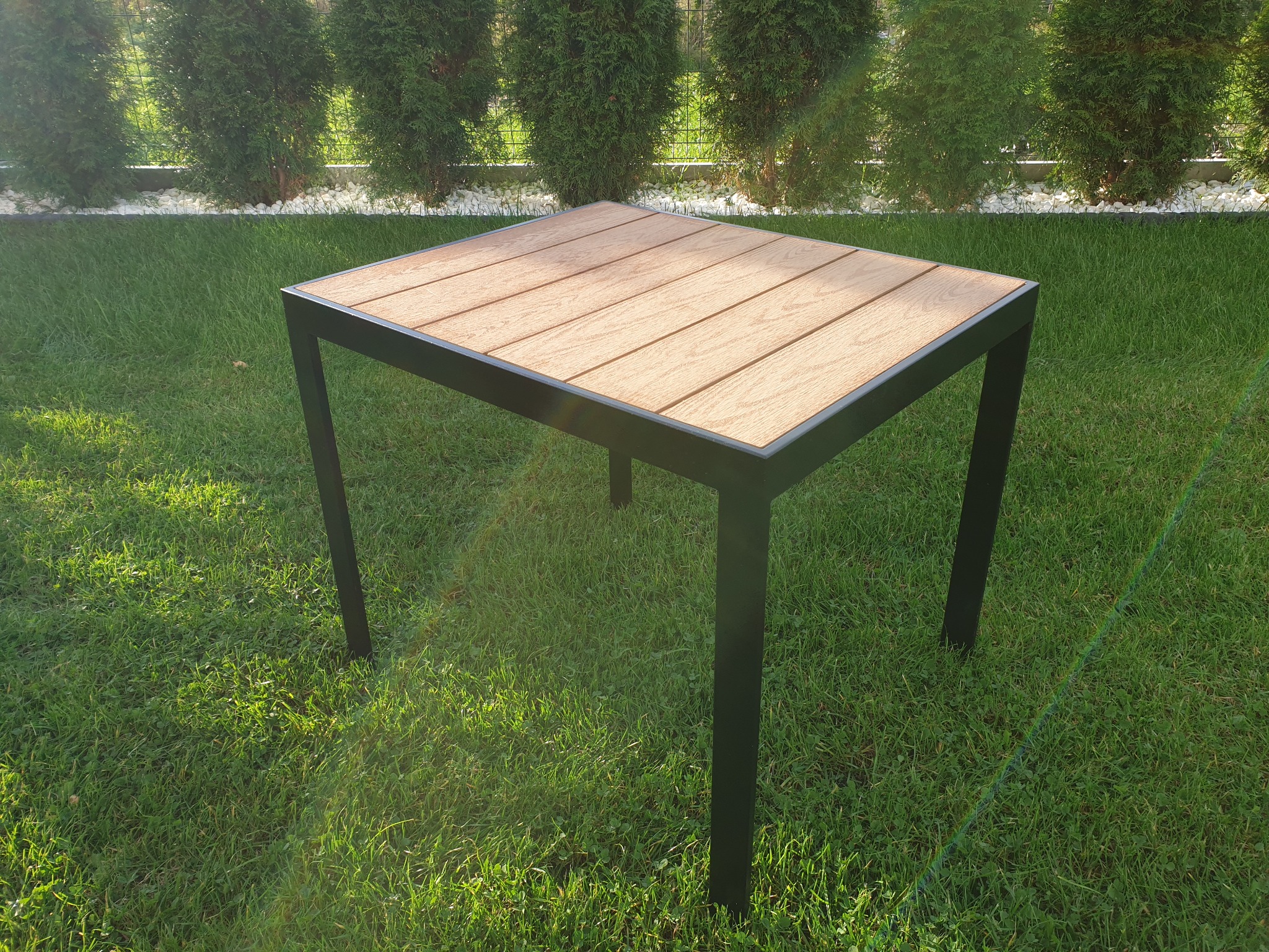 Stół ogrodowy z drewnopodobnym blatem i czarnymi, metalowymi nogami, ustawiony na trawie, w tle żywopłot.
