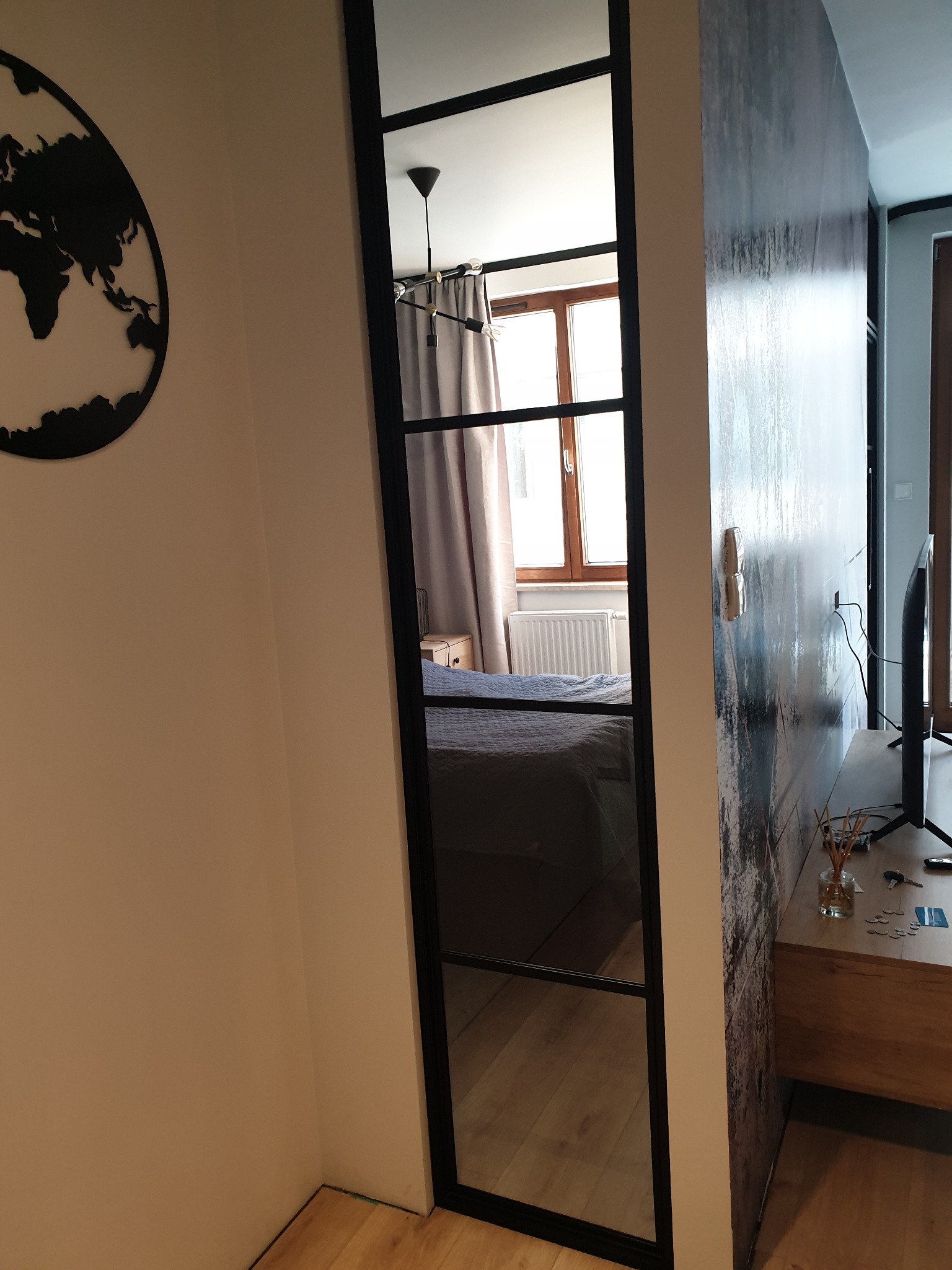 Przeszklone drzwi przesuwne z czarną ramą, widok na sypialnię z łóżkiem i oknem, dekoracja ścienna z mapą świata i abstrakcyjny wzór na ścianie.