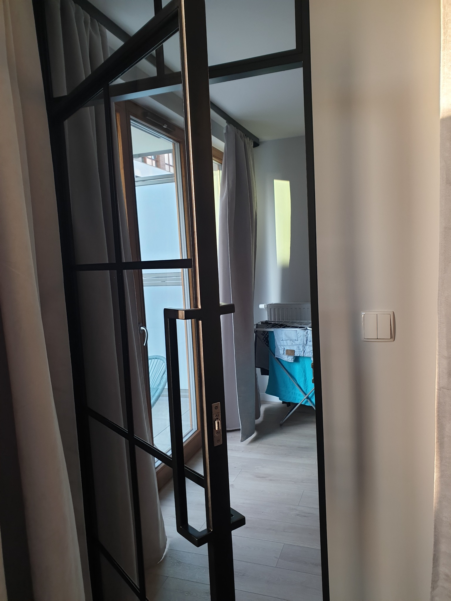 Częściowo otwarte, czarne, metalowe drzwi w stylu industrialnym z widokiem na balkon i suszarkę na pranie w tle.