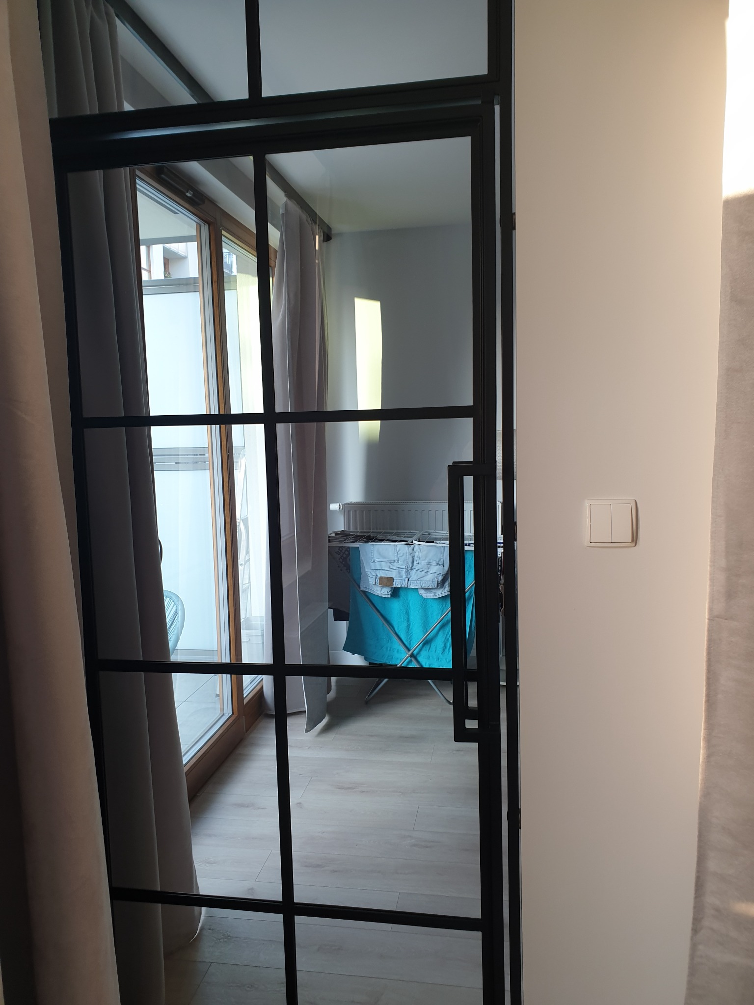 Czarna, stalowa konstrukcja drzwi z szybami w stylu loftowym, widoczna z boku, z fragmentem ściany z włącznikiem światła; w tle suszarka na pranie.