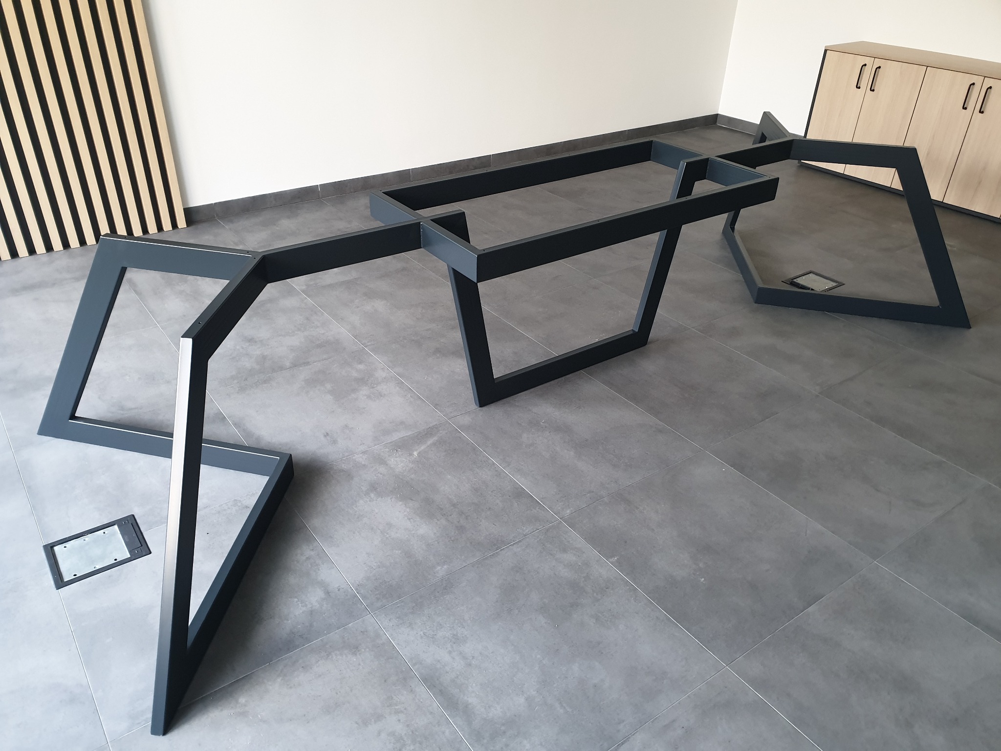 Metalowa konstrukcja stołu o geometrycznym kształcie, malowana na szaro, stoi na szarych płytkach w jasnym wnętrzu biurowym.