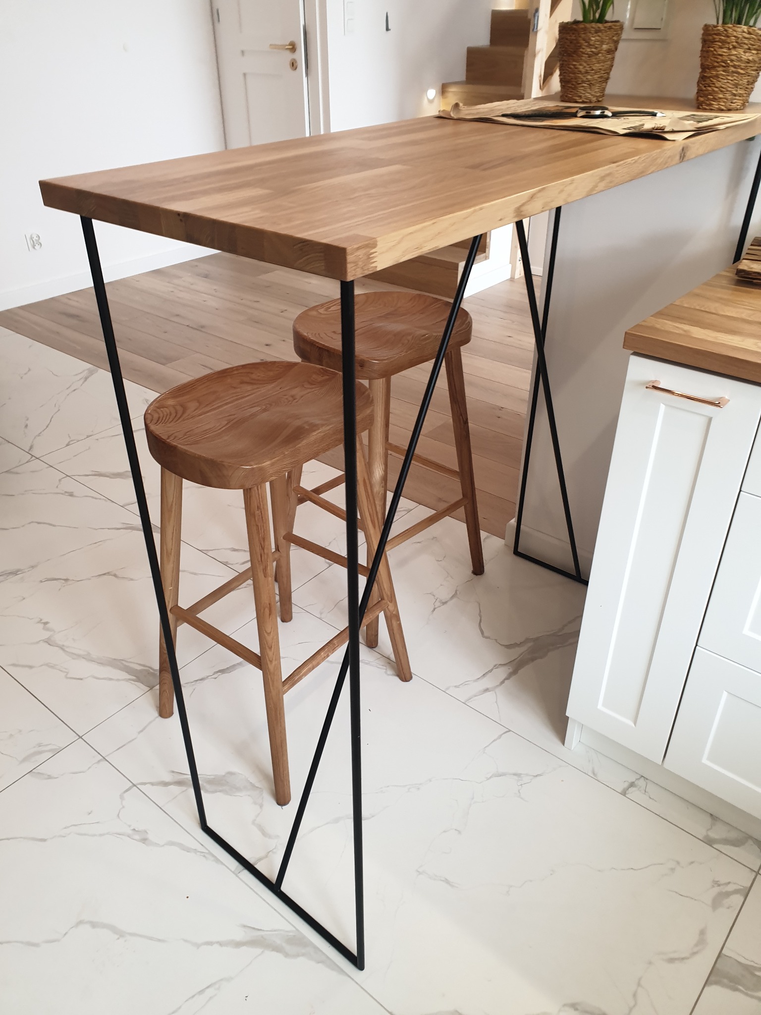 Blat dębowy stołu barowego na czarnych, spawanych nogach z dwoma drewnianymi stołkami, na tle białych szafek kuchennych i marmurowej podłogi.