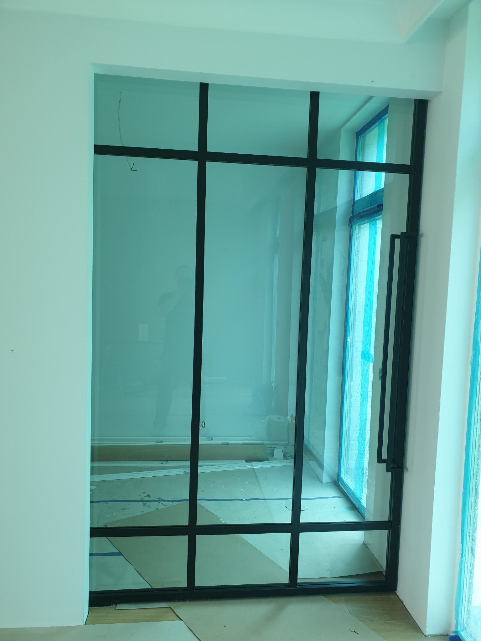 Nowoczesne, przeszklone drzwi z czarnymi szprosami, widoczne w trakcie montażu, zabezpieczone folią okna w tle.