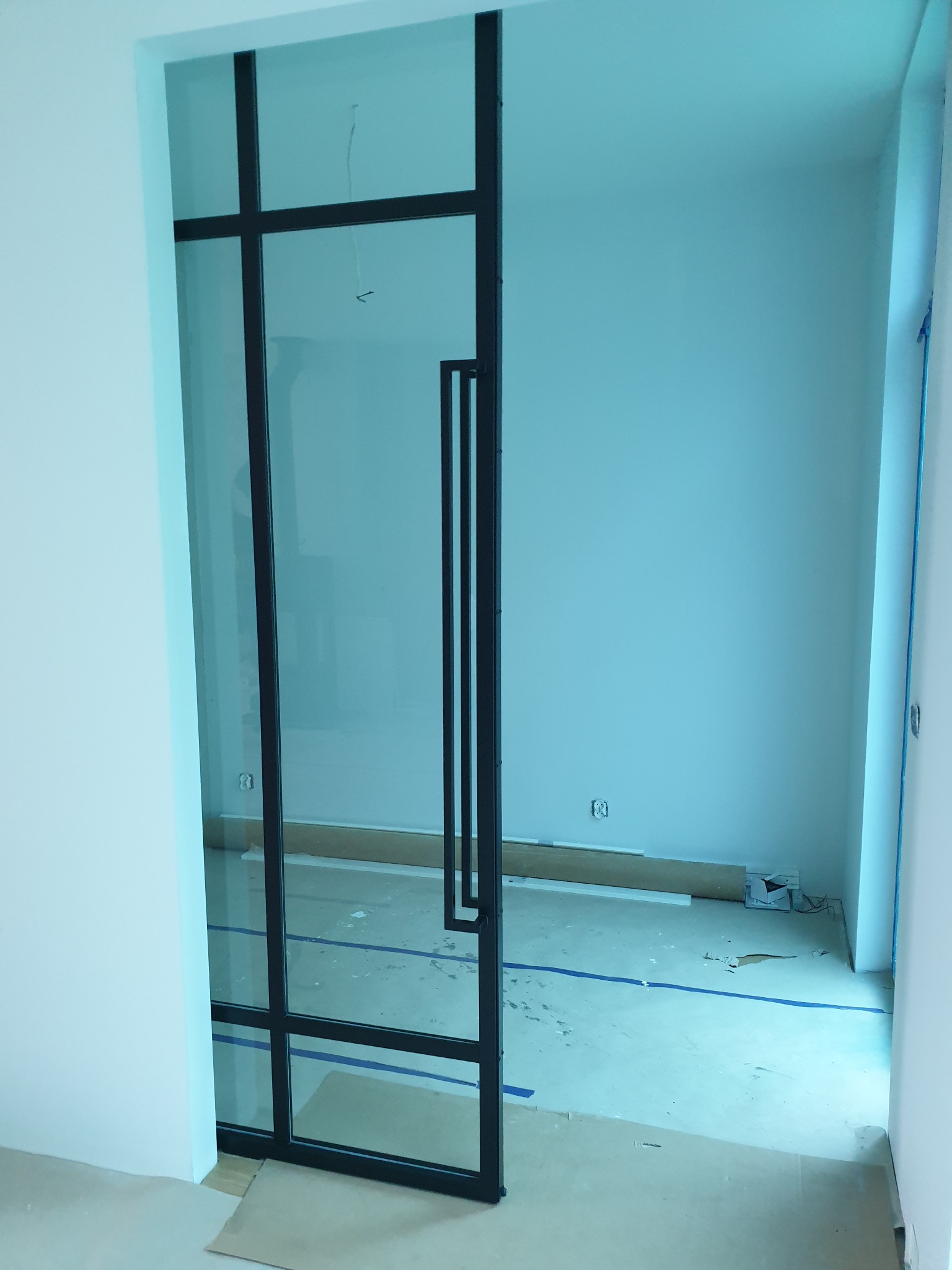 Szklane drzwi przesuwne z czarnymi metalowymi szprosami i długim uchwytem, widok na wnętrze w trakcie remontu, zabezpieczona podłoga.