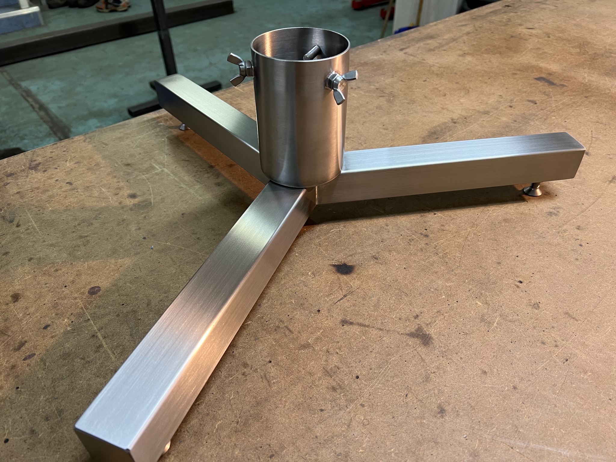 Metalowa podstawa z trzema ramionami i centralnym cylindrem, połączona śrubami motylkowymi, leżąca na drewnianym blacie.