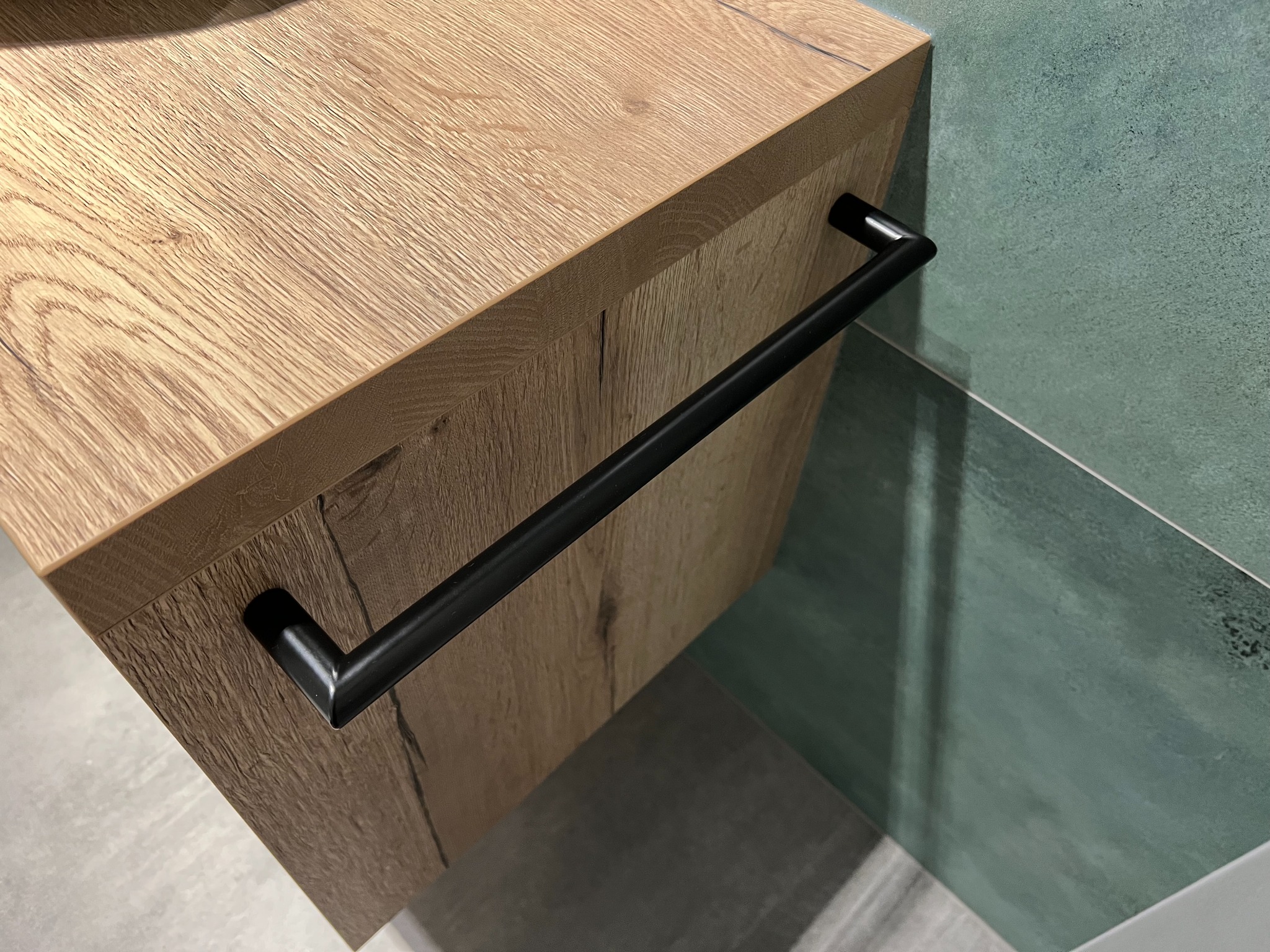 Fragment szafki łazienkowej z drewnopodobnym frontem i czarnym, minimalistycznym uchwytem na tle turkusowych płytek.