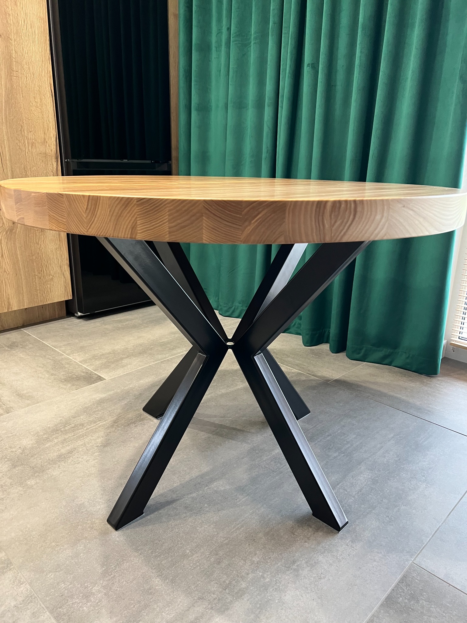 Okrągły stół z drewnianym blatem o wyraźnym usłojeniu i czarnymi, metalowymi nogami w kształcie litery X, na tle zielonej zasłony i jasnej podłogi z płytek.