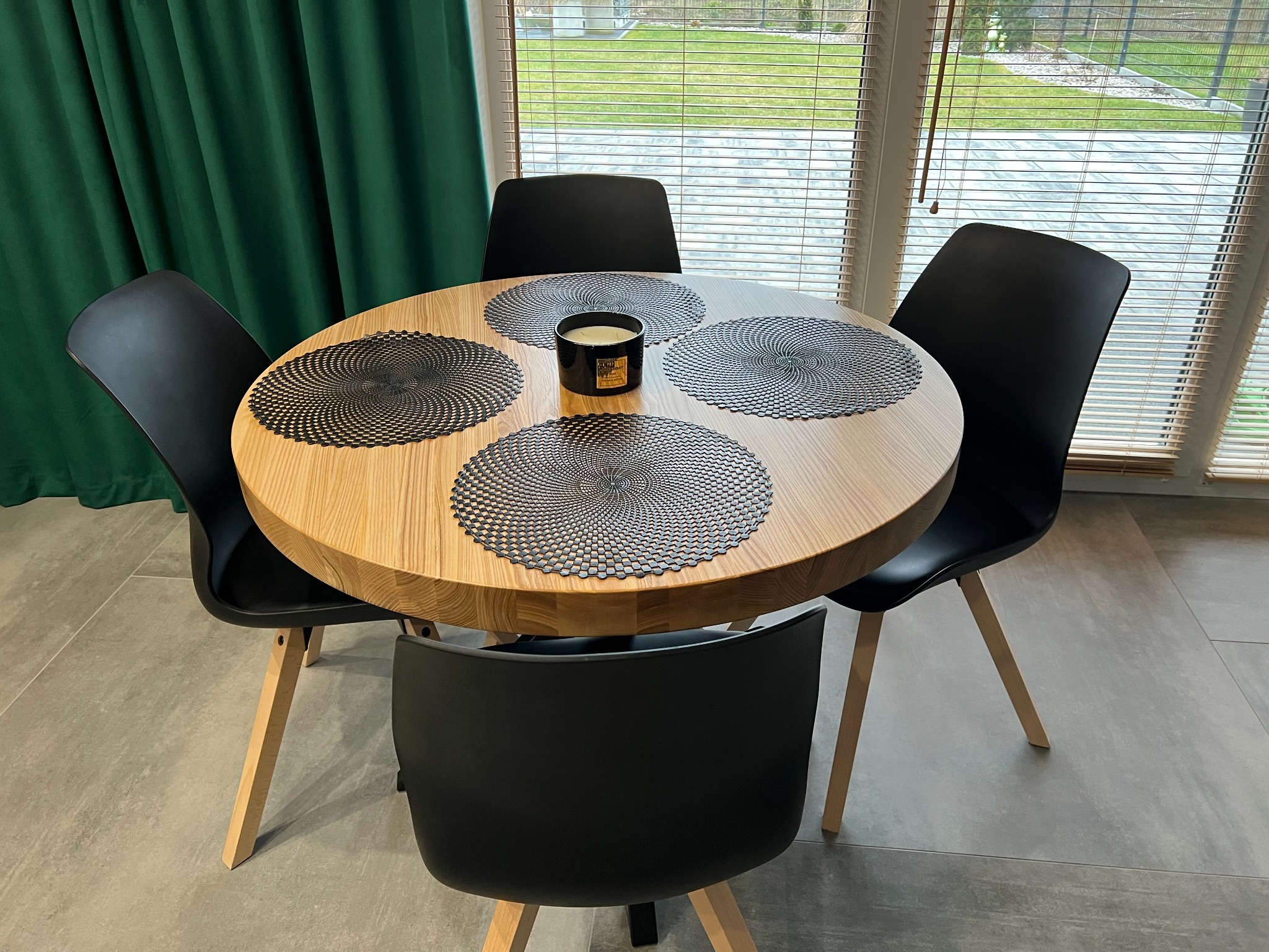 Okrągły, drewniany stół z czterema czarnymi krzesłami, ustawiony na szarych płytkach podłogowych, z dekoracyjnymi podkładkami i czarną świecą na środku, widok zza okna z żaluzjami.