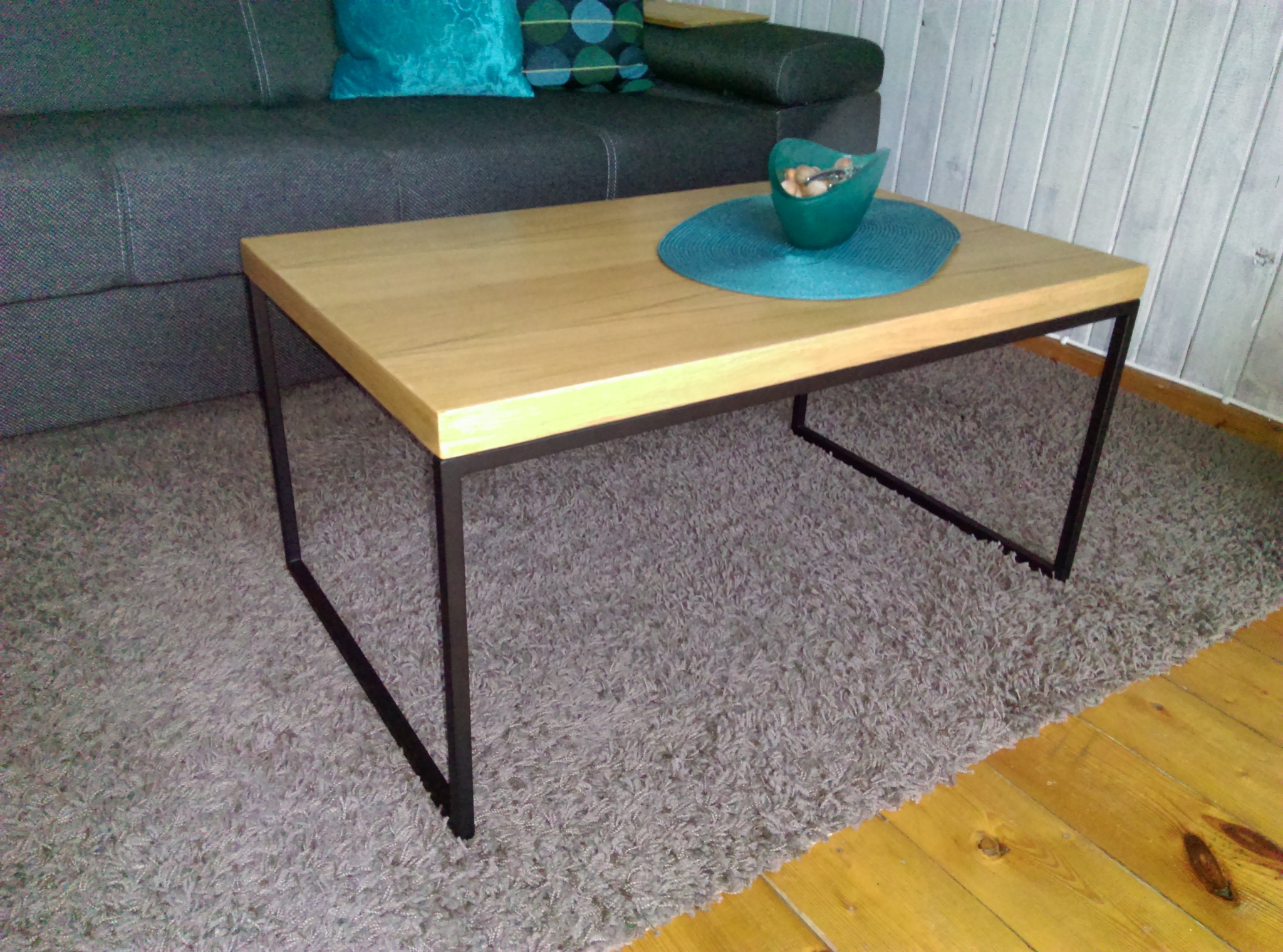 Stolik kawowy z jasnym drewnianym blatem i czarnymi metalowymi nogami, ustawiony na puszystym dywanie w salonie, z turkusową miseczką i podkładką na blacie, obok szarej sofy z turkusowymi poduszkami.
