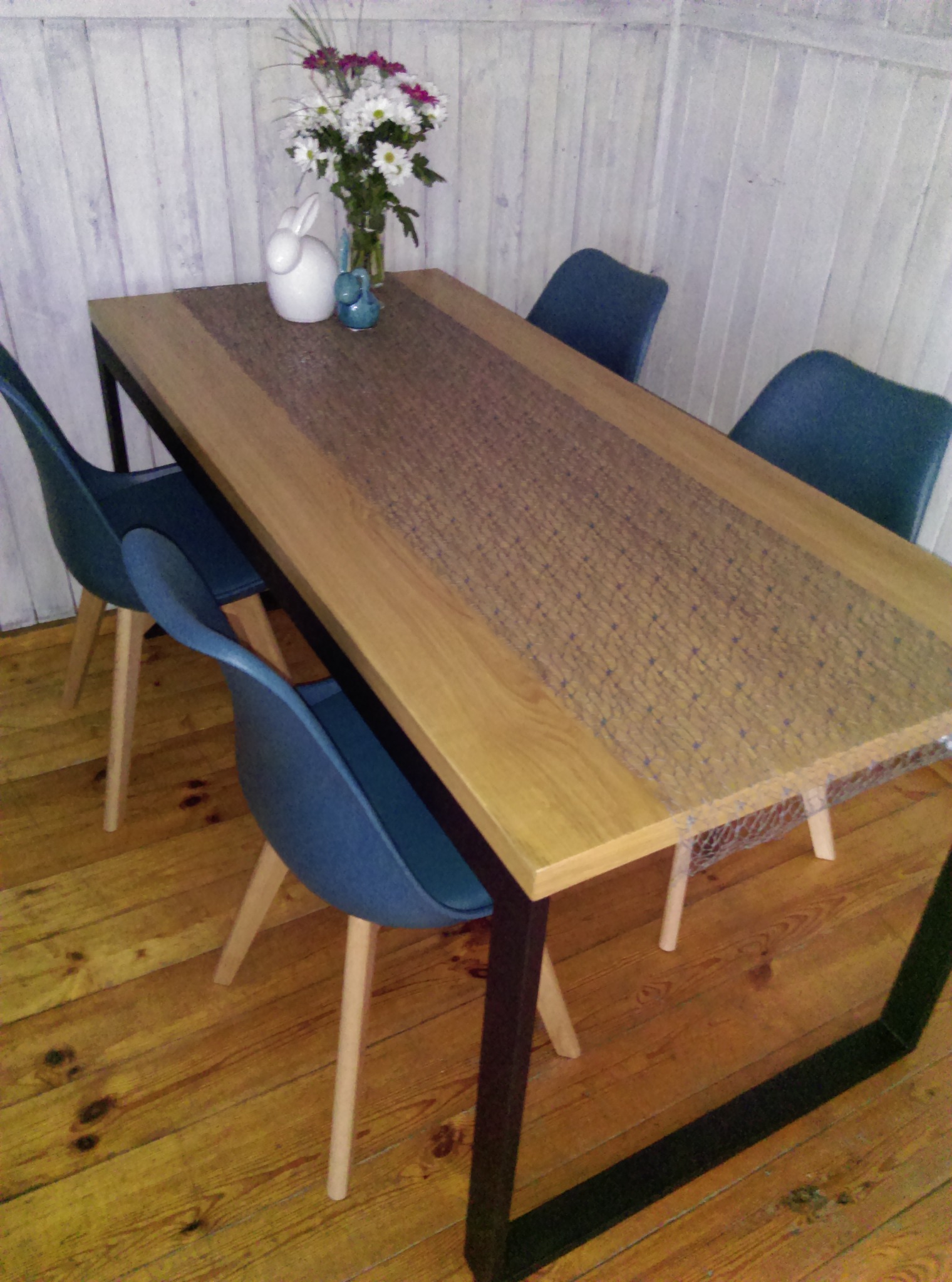 Stół z drewnianym blatem i metalową podstawą, nakryty siatkową bieżnikiem, otoczony niebieskimi krzesłami, z wazonem kwiatów i ceramiczną figurką królika. Podłoga drewniana, ściana z paneli...