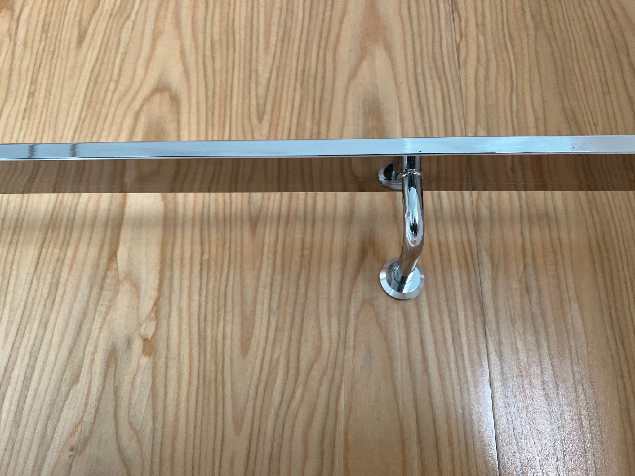 Chromowana poręcz zamontowana na jasnej, drewnianej ścianie, widoczne mocowanie i odbicie światła na metalu.