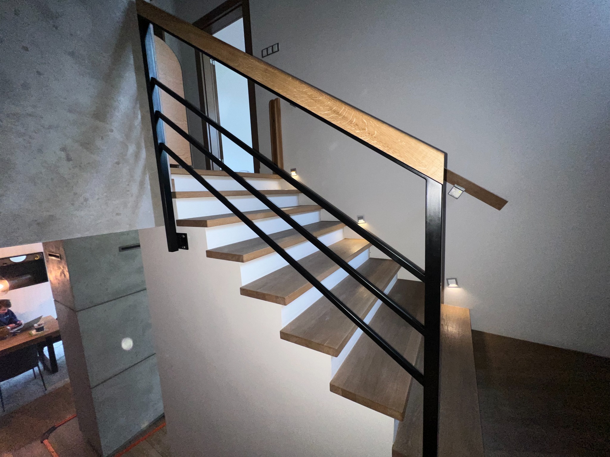 Nowoczesne schody drewniane z czarną metalową balustradą i subtelnym oświetleniem punktowym, widoczne z dołu, z fragmentem ściany o surowej fakturze w tle.