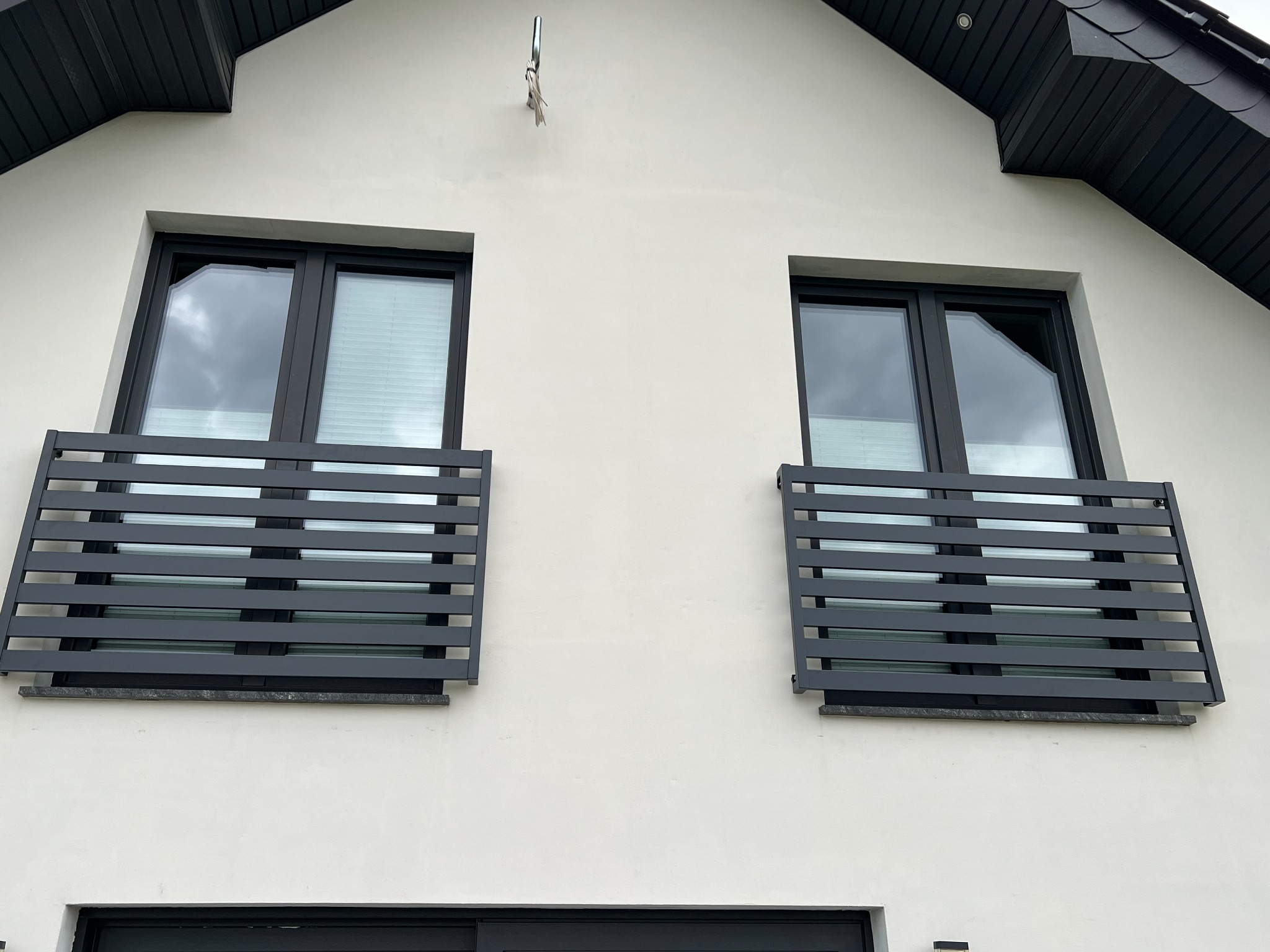 Elewacja domu z dwoma oknami zabezpieczonymi poziomymi, szarymi, metalowymi barierkami. Widoczne czarne ramy okienne i biała ściana budynku.