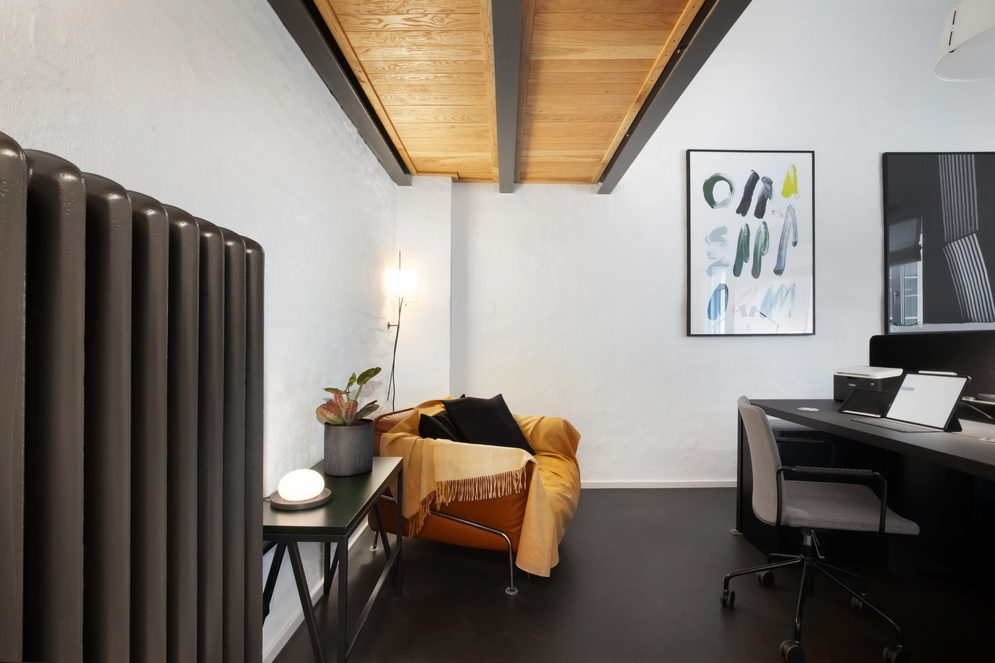 Nowoczesne wnętrze biurowe z ciemną podłogą, grzejnikiem żeberkowym, drewnianym sufitem, fotelem, biurkiem i abstrakcyjnymi obrazami na ścianie.