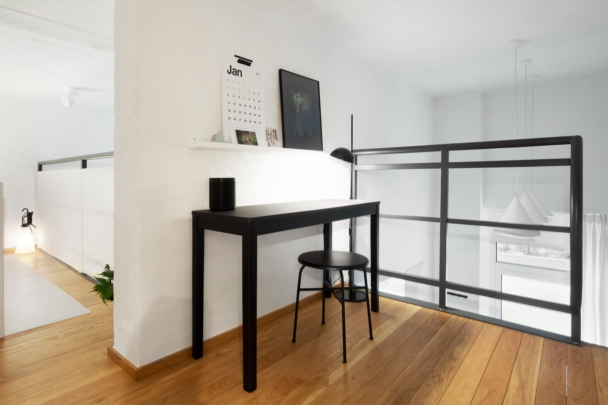Minimalistyczne wnętrze z czarną metalową balustradą, biurkiem i stołkiem na drewnianej podłodze, kalendarz na ścianie.