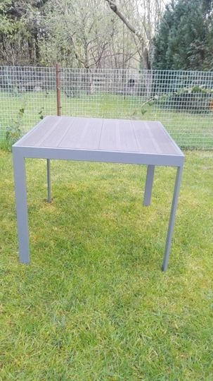 Kwadratowy stół ogrodowy z szarym blatem imitującym drewno i metalową ramą, ustawiony na trawniku, z siatką ogrodzeniową w tle.