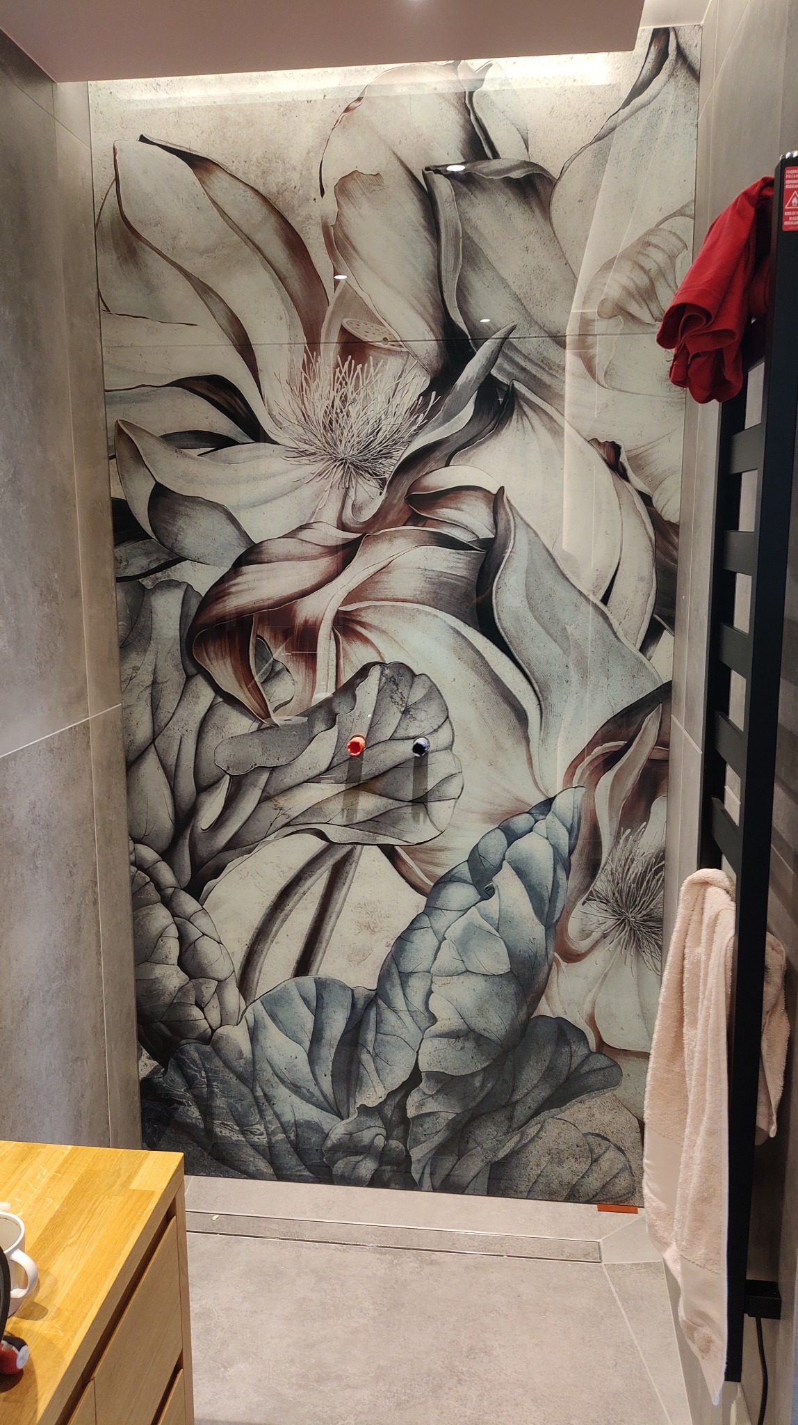 Szklany panel prysznicowy z nadrukiem UV przedstawiającym motyw kwiatowy, przymocowany do ściany za pomocą dwóch przyssawek z czerwonym i niebieskim uchwytem. W tle widoczny grzejnik drabinkowy...