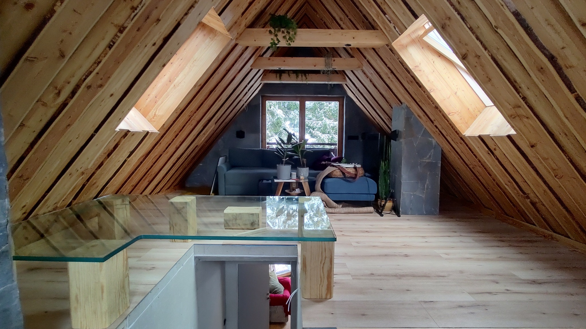 Szklany podest na drewnianych wspornikach w adaptowanym poddaszu z widocznymi elementami konstrukcji dachu i oknami dachowymi, w tle część wypoczynkowa z sofą.