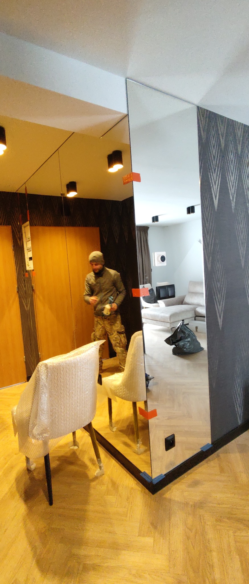 Montaż lustra na ścianie z wzorzystą tapetą, widoczny pracownik w odzieży roboczej, zabezpieczone krzesła i elementy mocujące.