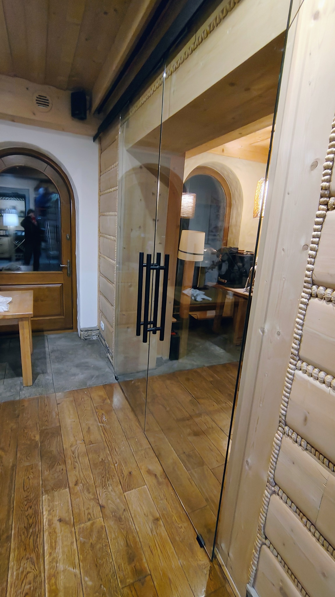 Przesuwne szklane drzwi z czarnym, minimalistycznym uchwytem w rustykalnym wnętrzu z drewnianą podłogą i ścianami zdobionymi w stylu góralskim.