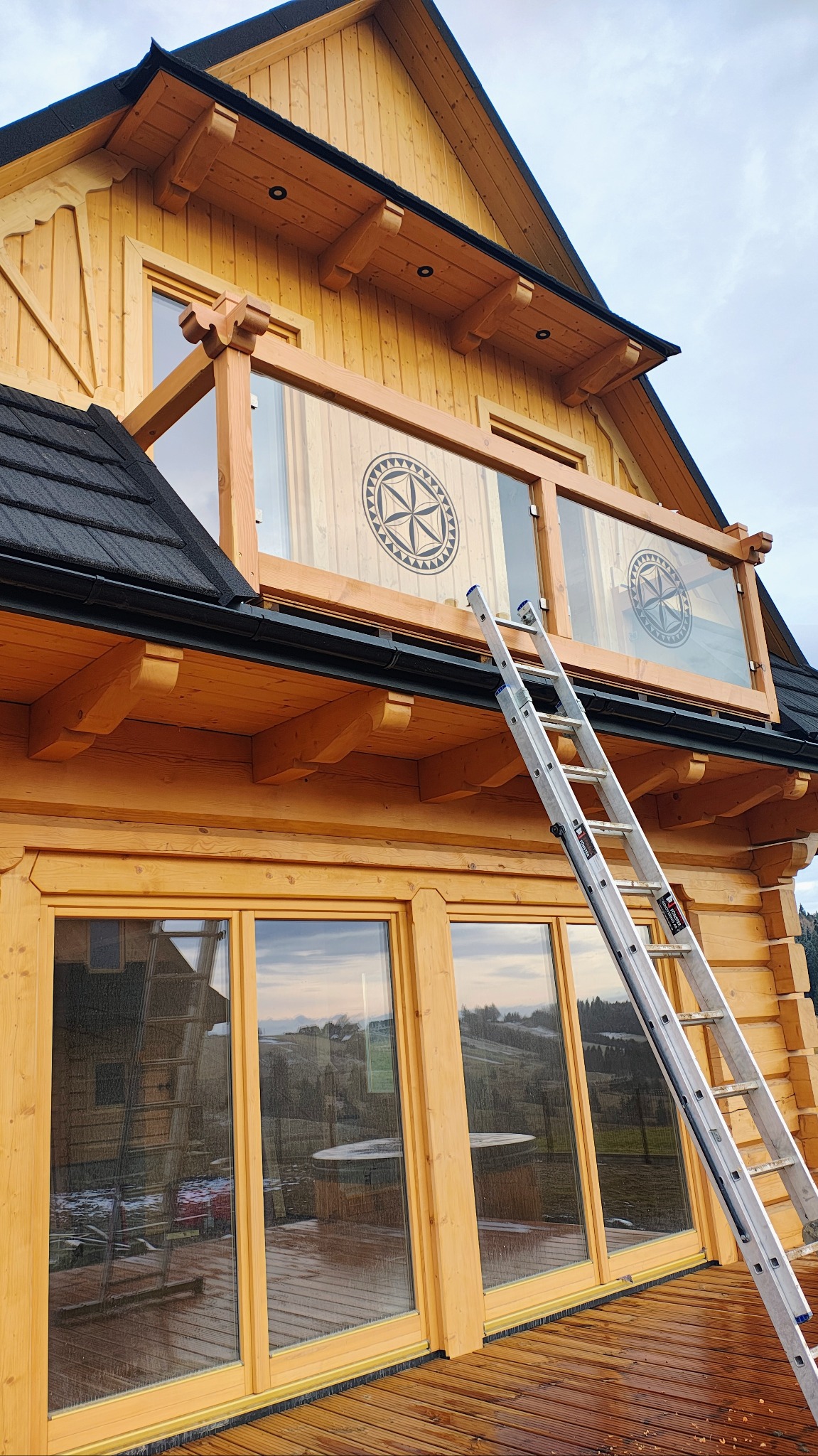 Balustrada balkonowa z drewnianą ramą i szkłem z motywem regionalnym na tle drewnianego domu, drabina oparta o balustradę.