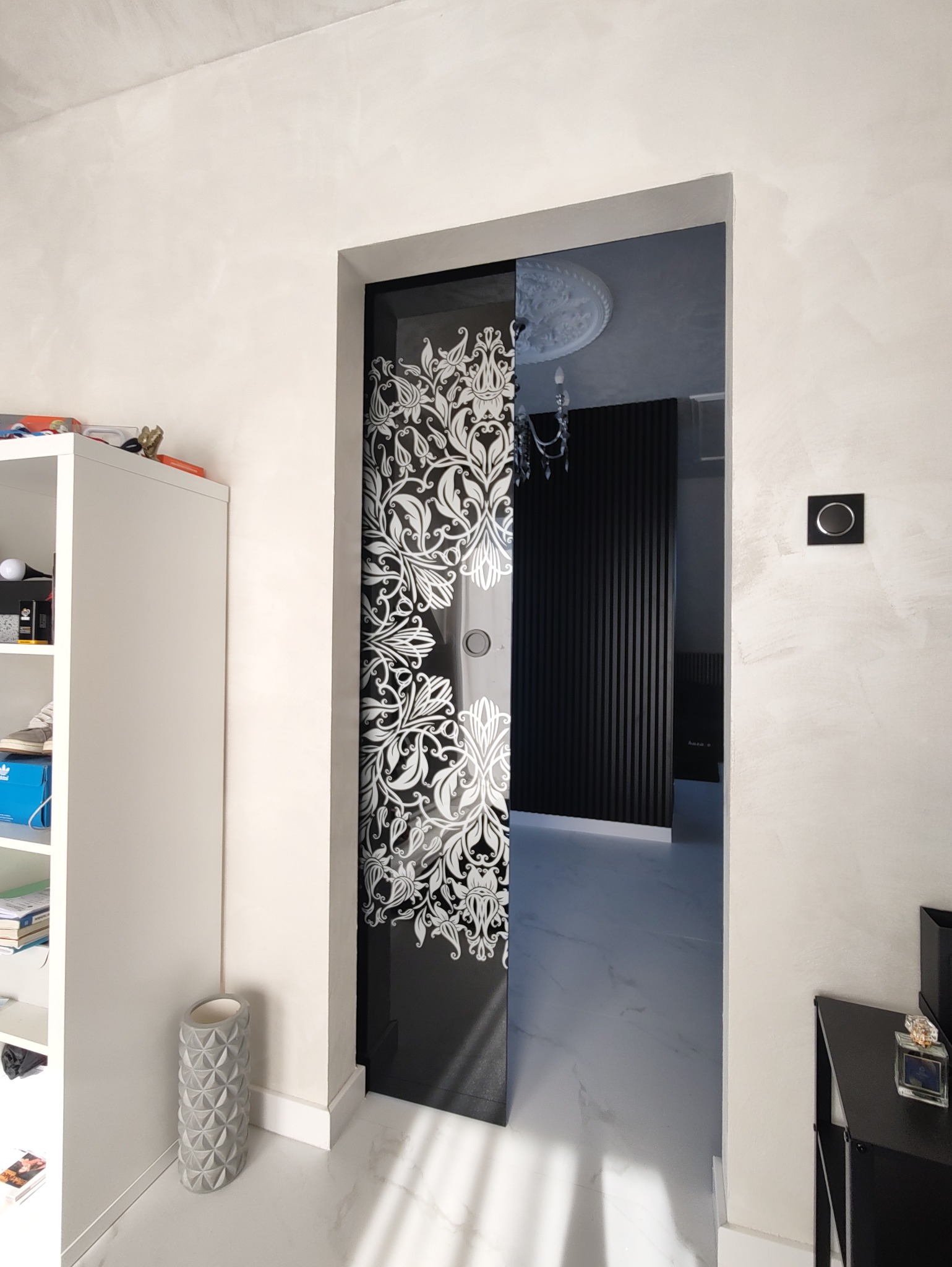 Szklane drzwi przesuwne z czarnym tłem i białym, roślinnym wzorem, w tle widoczny fragment pokoju z czarnymi panelami na ścianie.