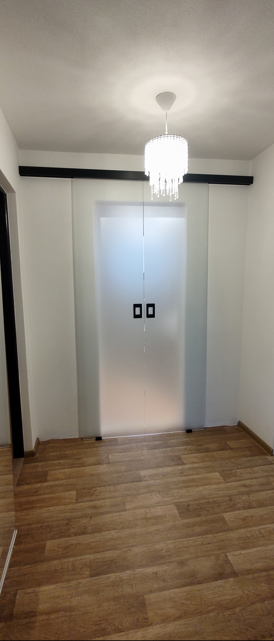 Nowoczesne, przesuwne drzwi dwuskrzydłowe ze szkła matowego z czarnymi, kwadratowymi uchwytami, zamontowane na czarnej szynie podsufitowej, oświetlone kryształowym żyrandolem.