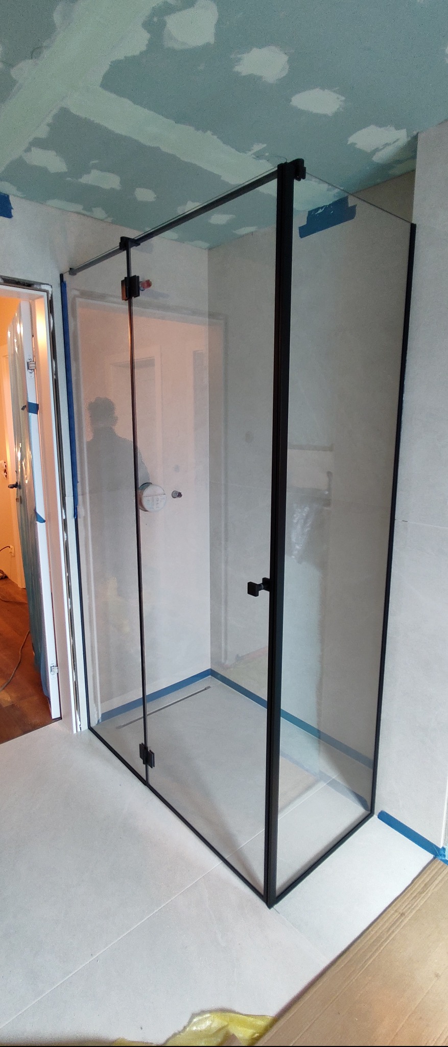 Kabina prysznicowa ze szkła z czarnymi profilami, widoczna w łazience w trakcie remontu, z taśmą malarską zabezpieczającą krawędzie.