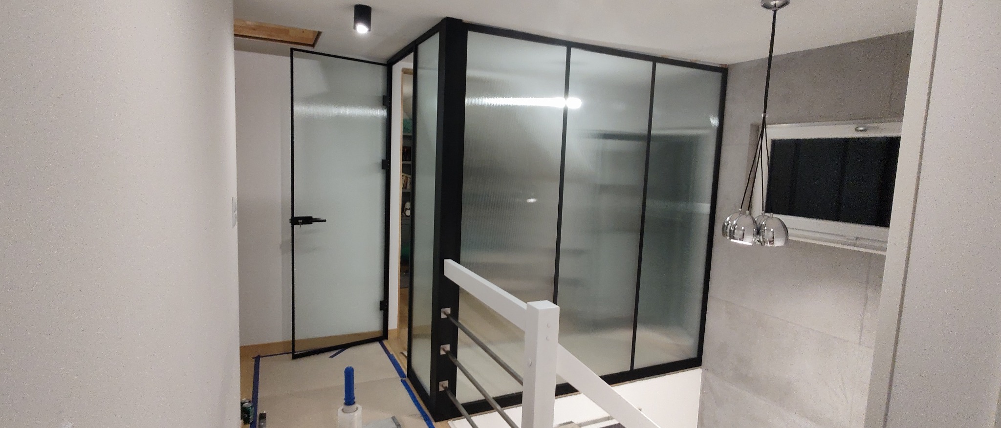 Szklana ścianka działowa z drzwiami w czarnej ościeżnicy aluminiowej, widoczna z perspektywy schodów z białą balustradą, w tle ryflowane szkło Visiosun.