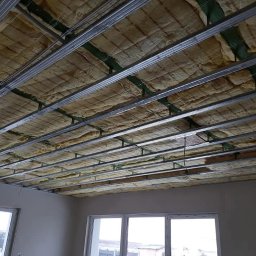 HYDRO PERFEKT Adam Kwaśny - Poddasze z izolacją z wełny mineralnej, widoczne metalowe profile konstrukcyjne sufitu podwieszanego, okna w tle. Etap prac wykończeniowych.