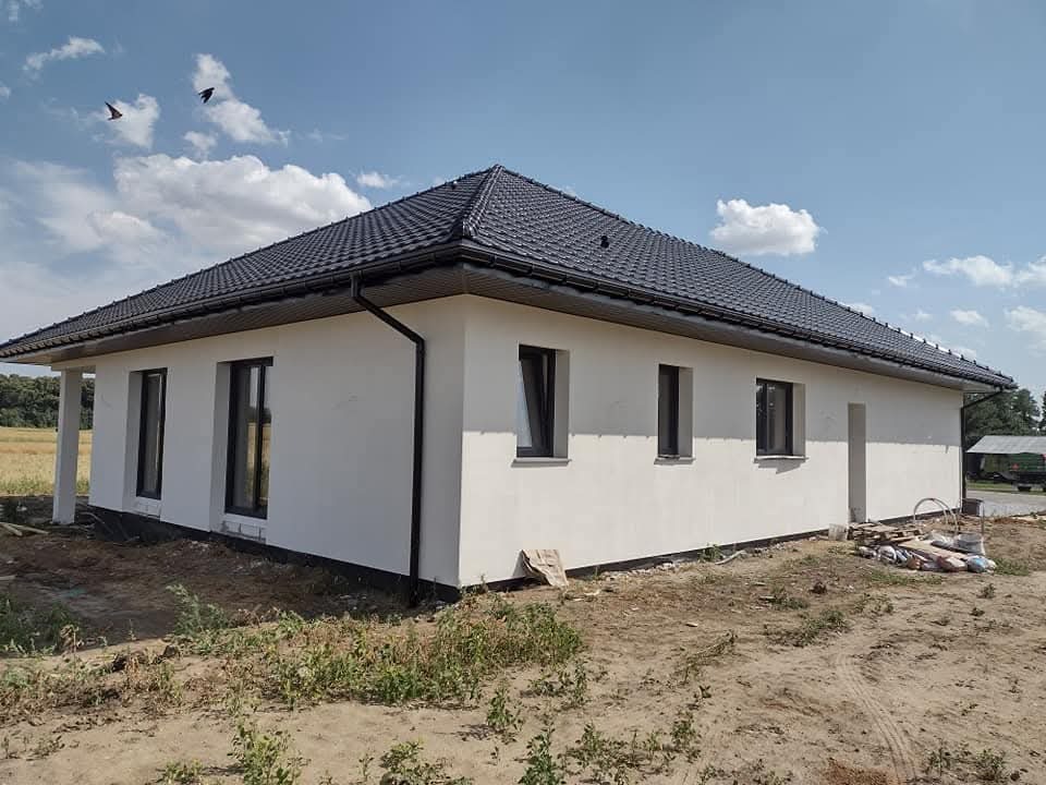 Biały dom z ciemnym dachem w słoneczny dzień. Widoczne okna, elewacja i fragment ogrodu. Dwa ptaki na niebie dodają dynamiki. Ujęcie z boku, podkreślające bryłę budynku.