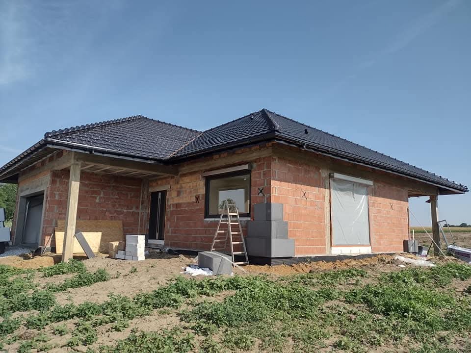 Surowy stan domu jednorodzinnego z czerwonej cegły, czarny dach, okna i drzwi wstawione, drabina oparta o ścianę, widok na budowę w słoneczny dzień.