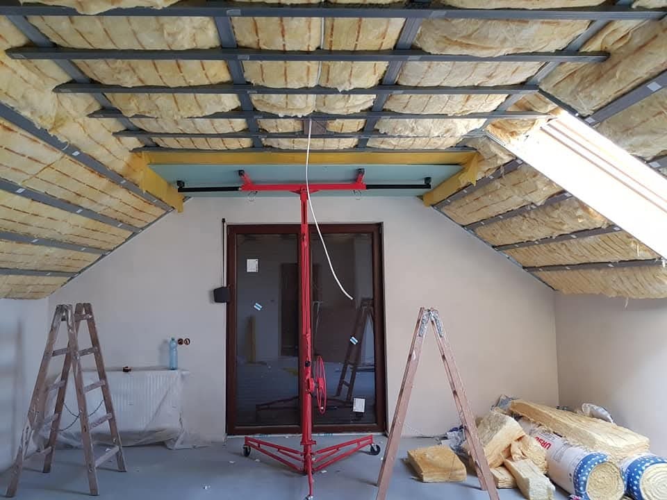 Poddasze w trakcie ocieplania wełną mineralną, widoczne metalowe profile, drabiny i podnośnik do płyt. Okno dachowe wpuszcza światło.