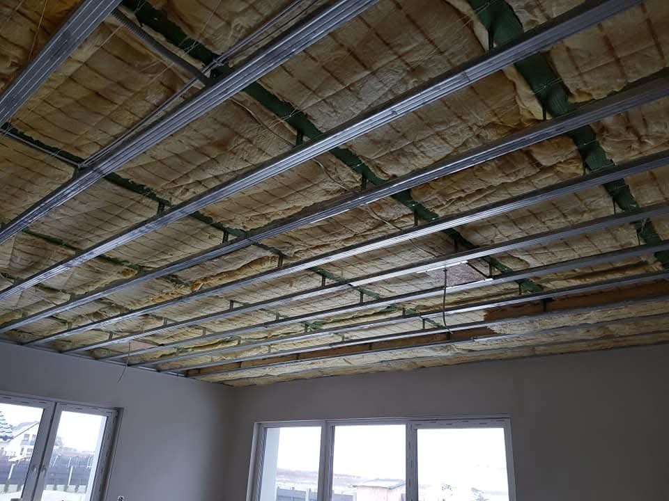 Poddasze z izolacją z wełny mineralnej, widoczne metalowe profile konstrukcyjne sufitu podwieszanego, okna w tle. Etap prac wykończeniowych.