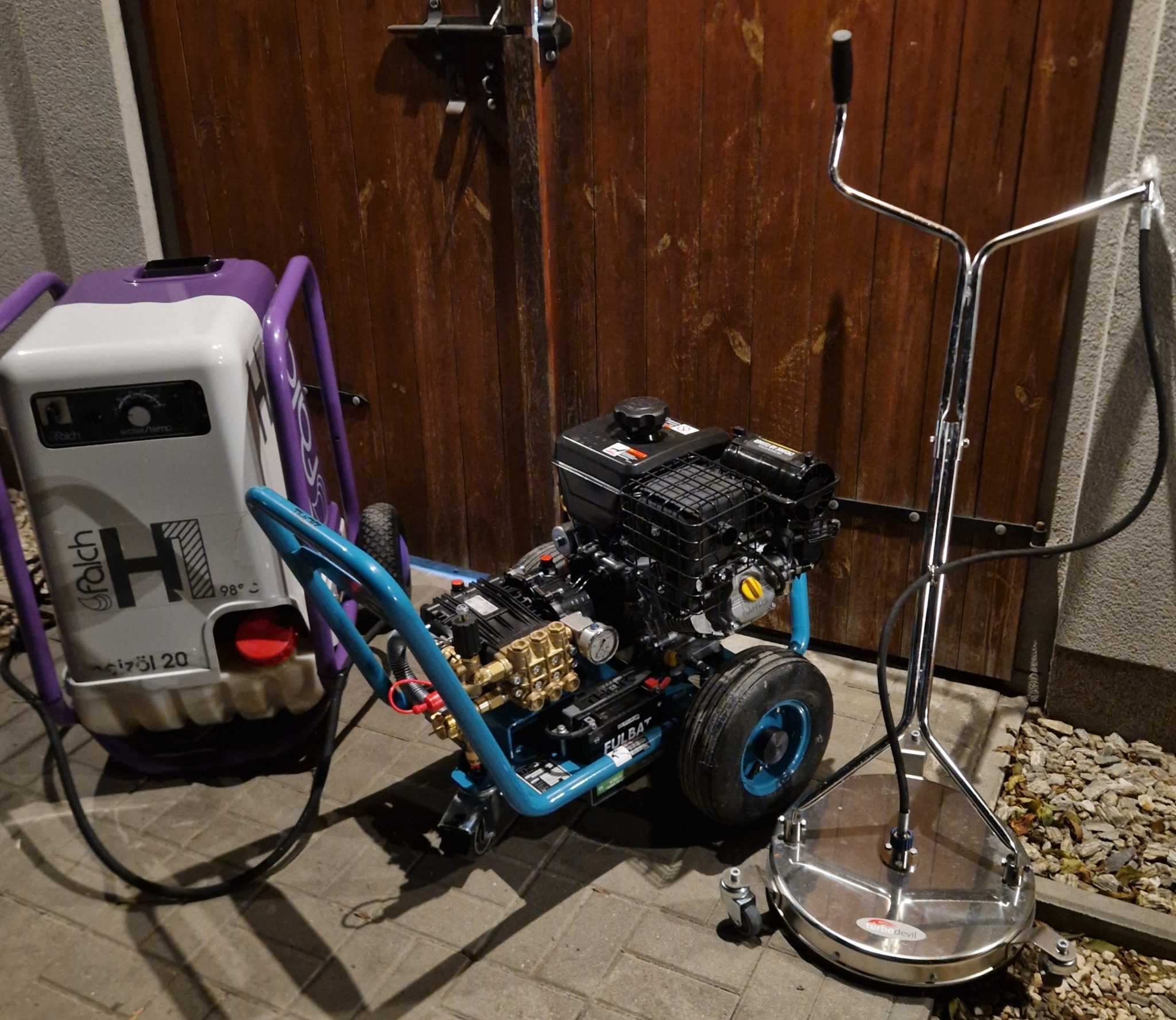 Profesjonalny sprzęt do mycia ciśnieniowego: myjka Falch i urządzenie do czyszczenia powierzchni Turbo Devil, gotowe do pracy na tle drewnianej bramy.