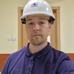 Mężczyzna w białym kasku ochronnym z logo firmy, ubrany w fioletową koszulkę polo, na tle ściany i drzwi. Widoczny włącznik światła.