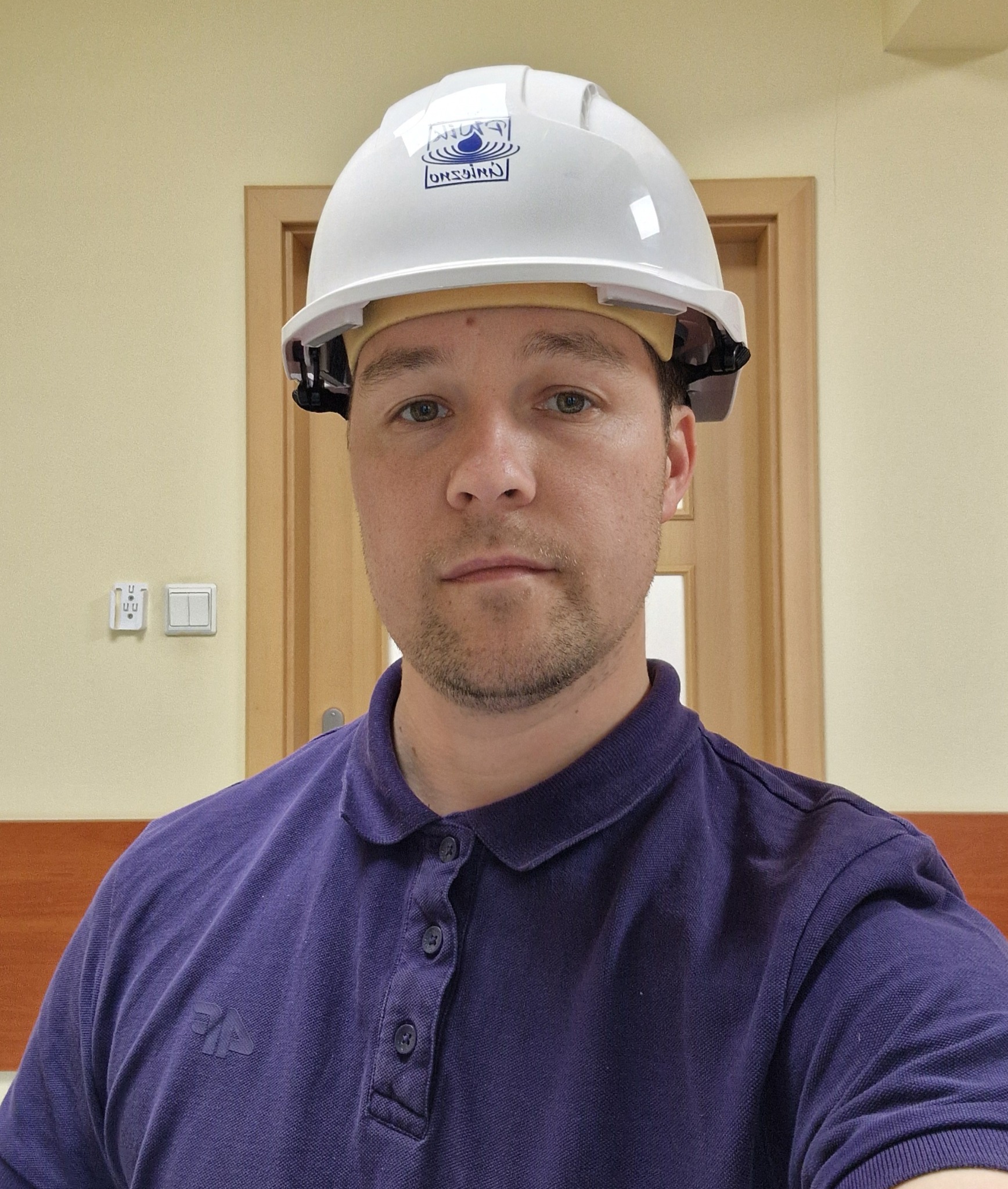 Mężczyzna w białym kasku ochronnym z logo firmy, ubrany w fioletową koszulkę polo, na tle ściany i drzwi. Widoczny włącznik światła.