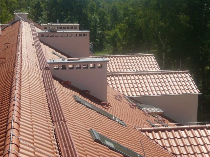 Czerwona dachówka ceramiczna na kilku połączonych dachach z kominami i oknami dachowymi, widok z góry na tle zieleni.