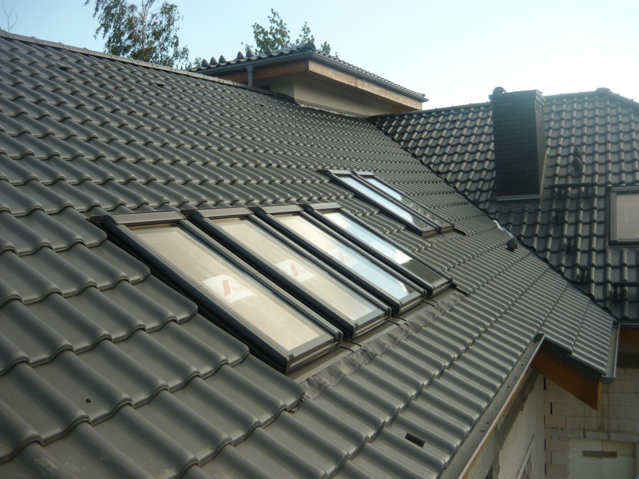 Czarny dach z dachówki ceramicznej z zamontowanymi oknami dachowymi, widok z góry na budynek w trakcie budowy.