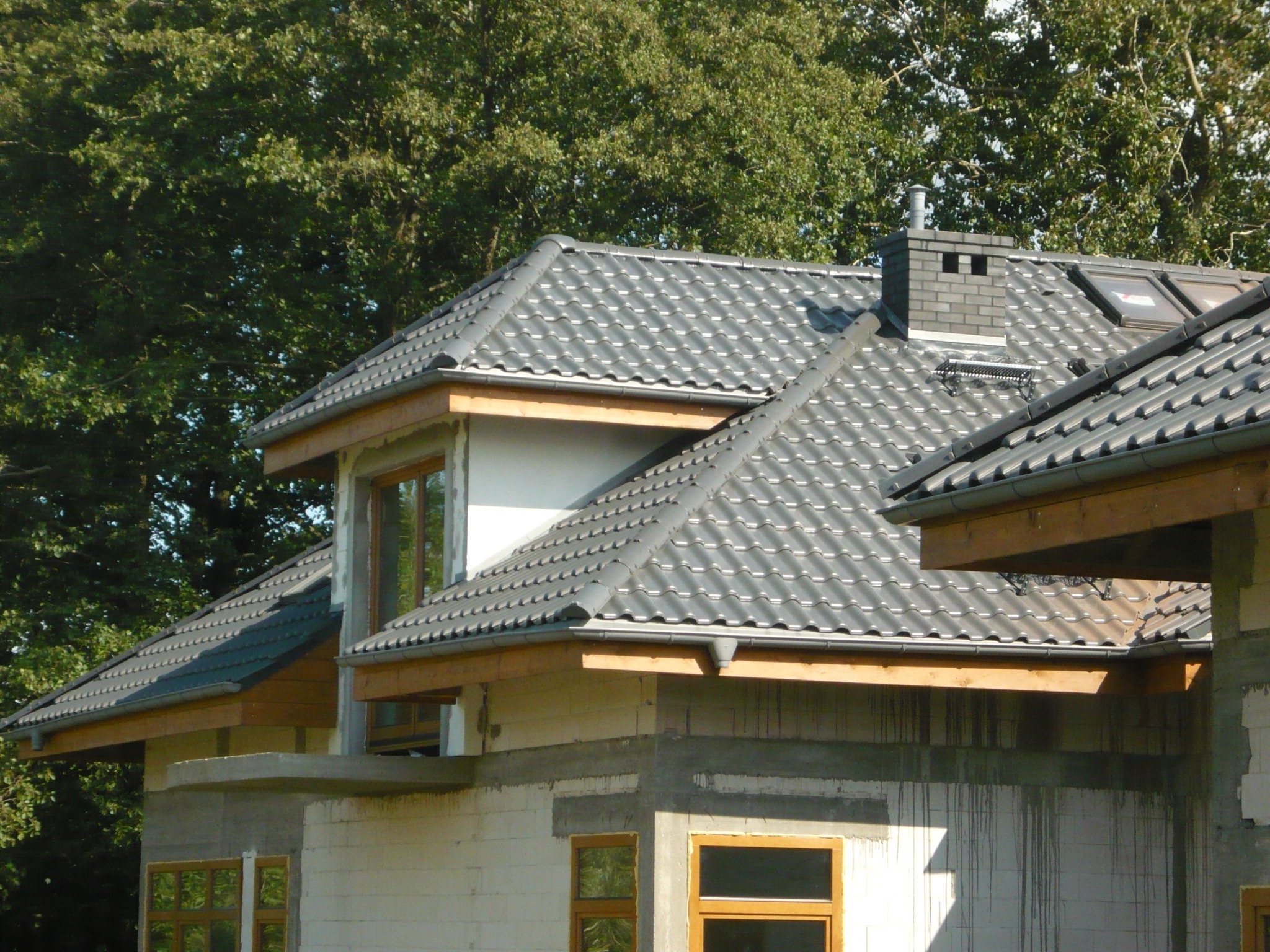 Nowy dom w trakcie budowy z grafitowym dachem z dachówki ceramicznej, widoczne okna dachowe, komin z cegły klinkierowej i orynnowanie.