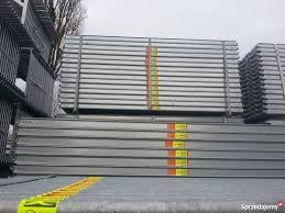 Stos metalowych, srebrnych rusztowań budowlanych z pomarańczowo-żółtymi etykietami, ułożonych równo na szarym podłożu, widok z boku.