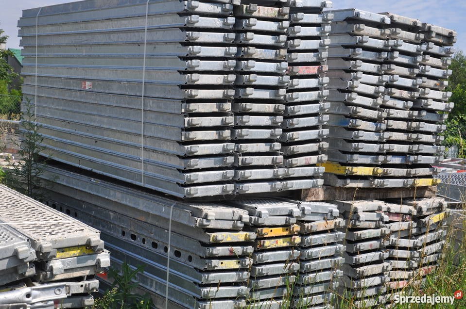 Stosy ocynkowanych, metalowych podestów rusztowaniowych ułożonych na paletach, widoczne ślady użytkowania i zabrudzenia.