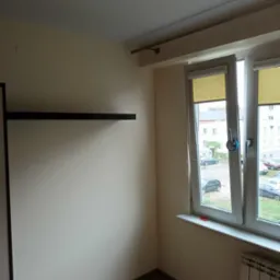 Pomalowane na beżowo wnętrze pokoju z drewnianą podłogą, grzejnikiem pod oknem z roletami i czarną półką na ścianie.