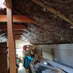 Poddasze ocieplone brązową wełną mineralną, widoczne drewniane belki konstrukcyjne, białe ściany z bloczków oraz kartony i różne przedmioty składowane w przestrzeni.