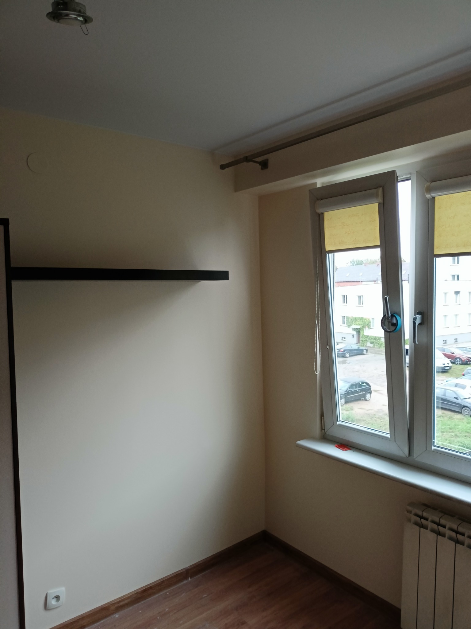 Pomalowane na beżowo wnętrze pokoju z drewnianą podłogą, grzejnikiem pod oknem z roletami i czarną półką na ścianie.