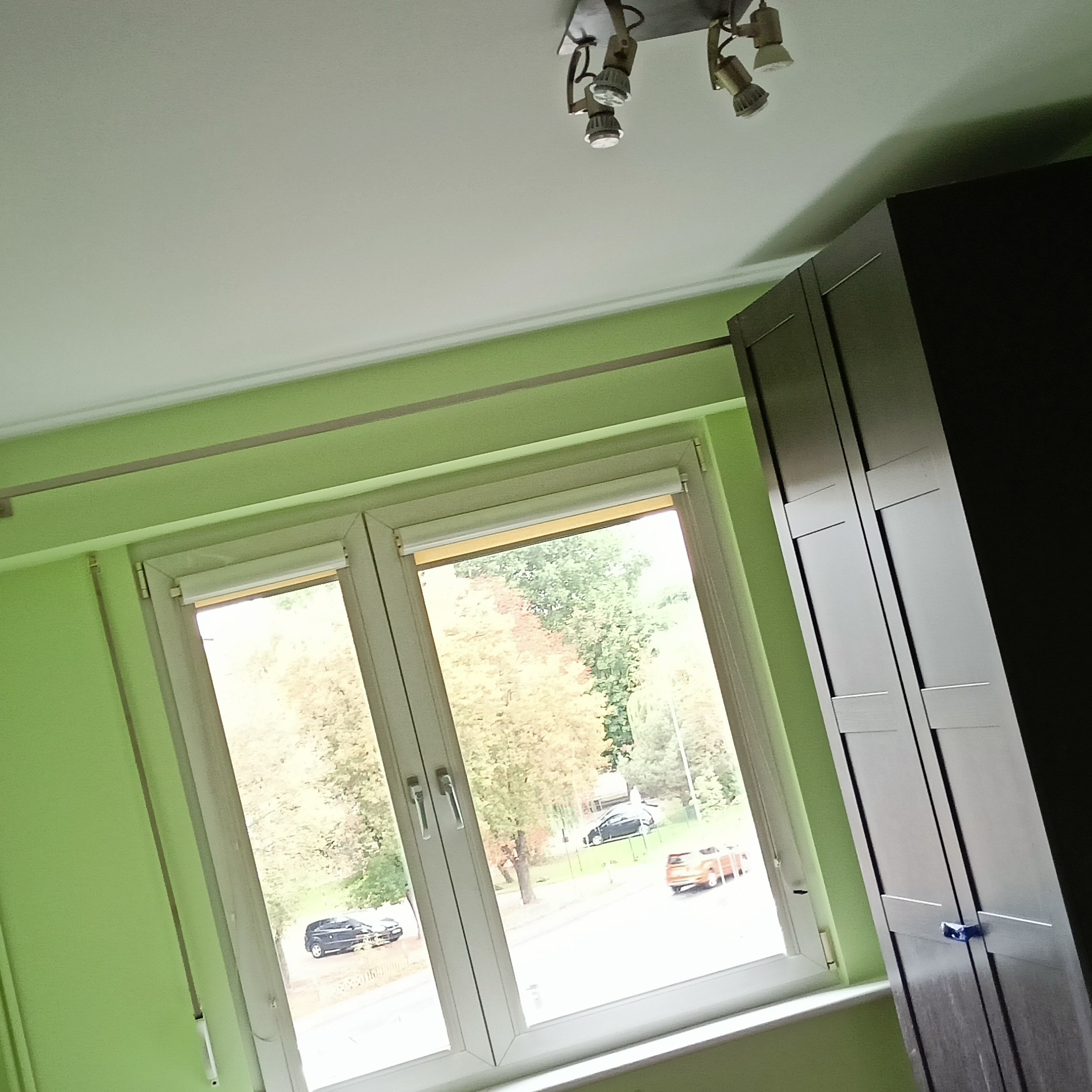 Jasnozielony pokój z oknem wychodzącym na ulicę z samochodami, widoczna ciemna szafa i lampa sufitowa z kilkoma reflektorami.