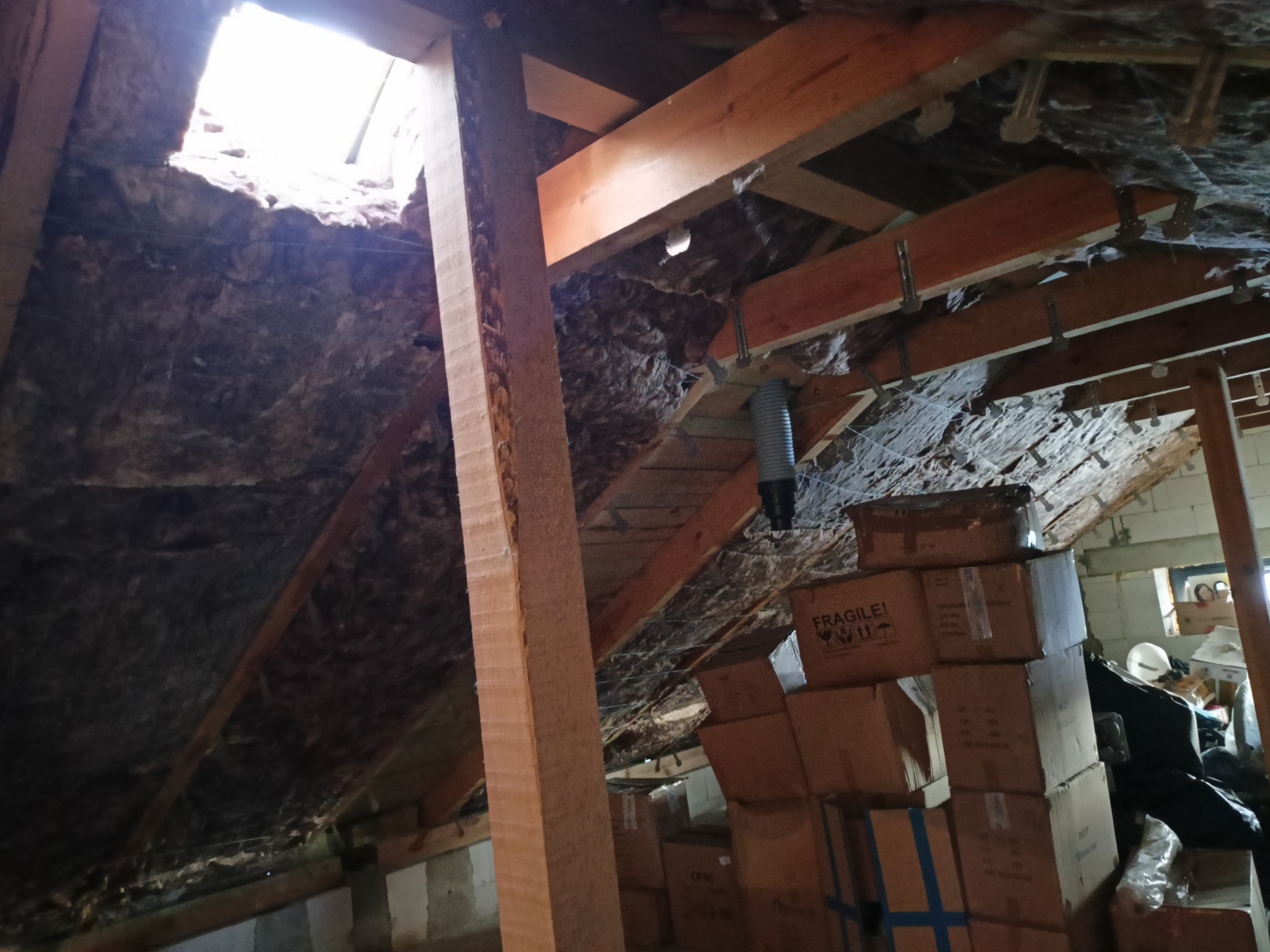 Poddasze w trakcie ocieplania wełną mineralną, widoczne drewniane krokwie, skrzynki z napisem 'FRAGILE' i fragment okna dachowego z wpadającym światłem.