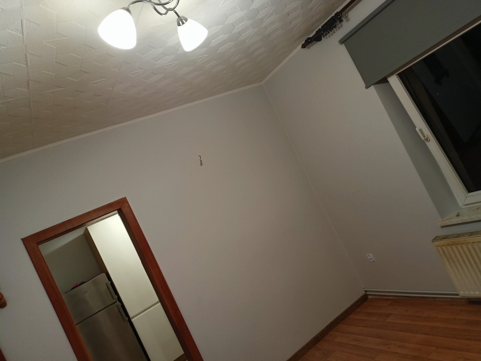 Pomalowane na biało wnętrze pokoju z drewnianą podłogą, widoczne okno z roletą, grzejnik, lampa sufitowa i fragment drzwi z widocznym wnętrzem szafy.