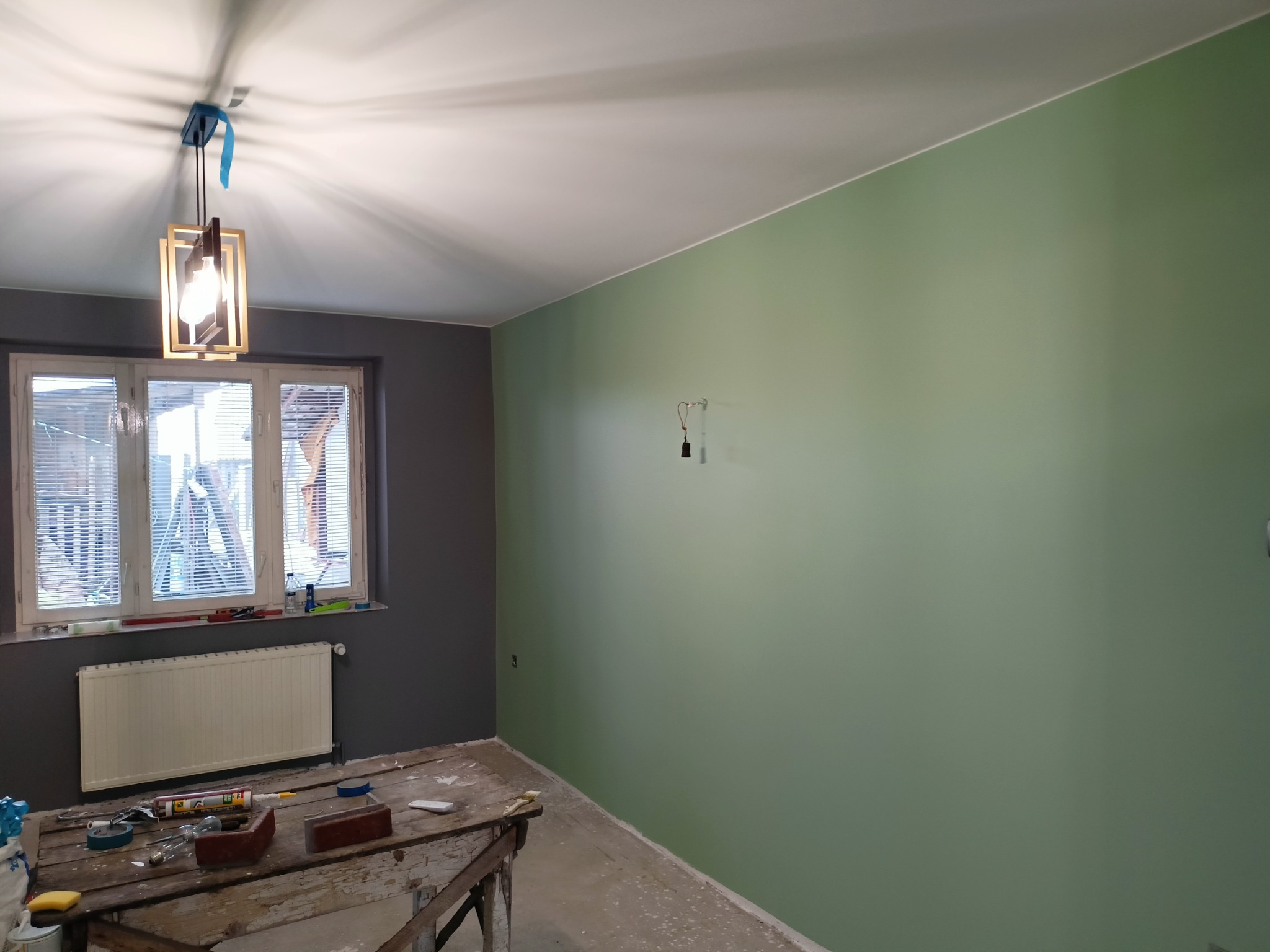 Pokój po malowaniu: ściana w kolorze pistacjowej zieleni, fragment szarej ściany przy oknie z grzejnikiem, drewniany stół roboczy z narzędziami i materiałami malarskimi, wisząca lampa...