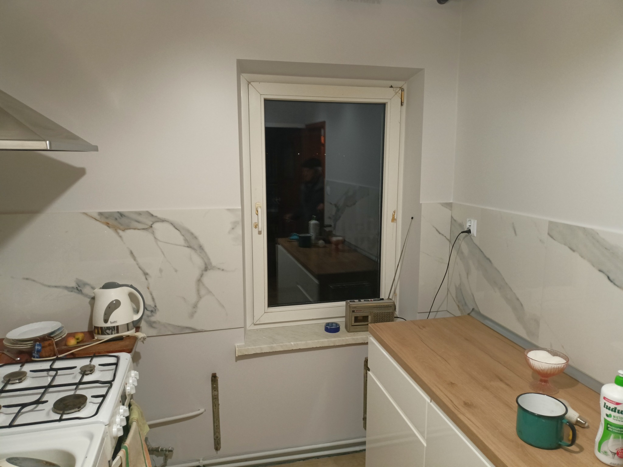 Kuchnia w trakcie prac wykończeniowych: marmurowe płytki na ścianie, widoczne gniazdko elektryczne, okno z radiem na parapecie, fragment kuchenki i blatu.